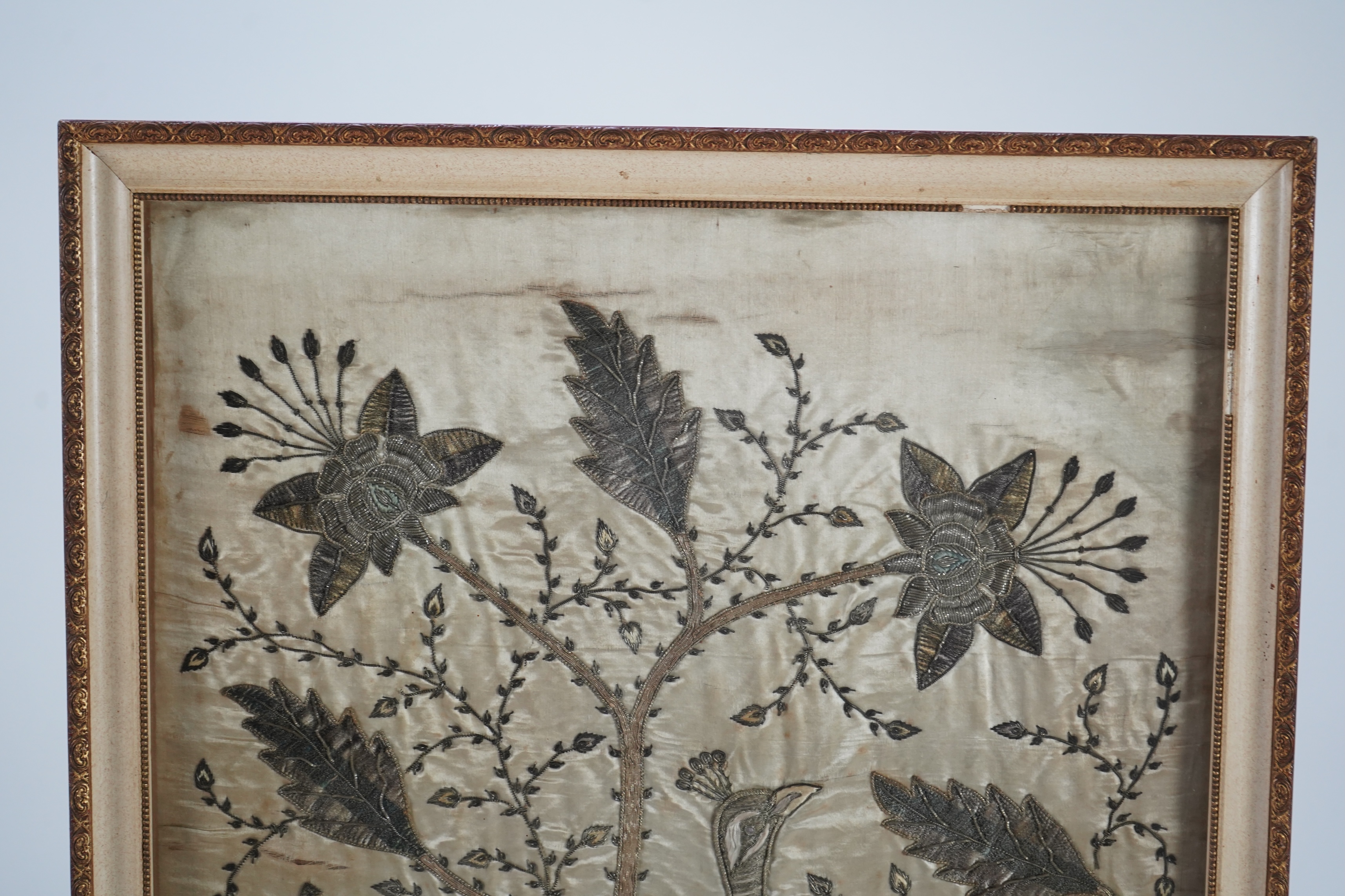 An Indian late 19th century silver metallic thread embroidery of a peacock in a flowering tree, embroidered on silk, parts of the embroidery worked in relief like stump work, 107cm x 52cm, framed. Condition - silk backin